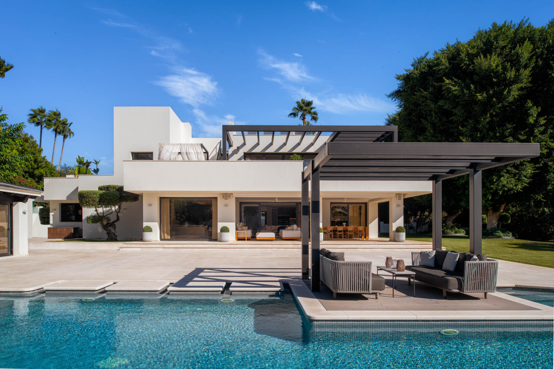 White modern villa facade seen across the swimming pool, with a pergola-shaded lounge on its own platform in the water.